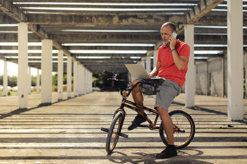 Naklejka premium Man siting on BMX bike holding notebook and talking on the phone on the street inside not ready building with striped shade from the sun 