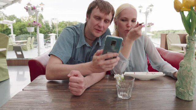 A young guy kisses a girl and takes a selfie while sitting in a restaurant. The girl at this time is eating ice cream and posing.