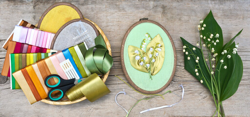 Needlework. Hand embroidery with satin ribbons of lily of the valley flowers in a frame on a old wooden bench in the garden and accessories for embroidery in a basket nearby. Flat lay, close-up
