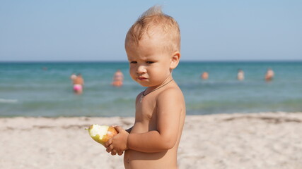 Cute child standing on the beach and eating a pear and looking displeased