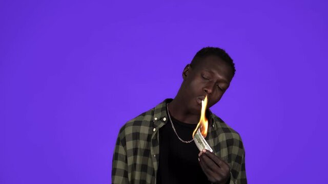 Serious man in casual clothes and golden chain on neck is lighting his cigarette with dollar note. Concept idea of wealth, rich men. Isolated over blue background