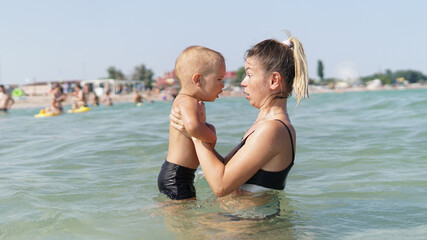 Fototapeta na wymiar Young mother standing in water in sea and holding smiling little son in her arms