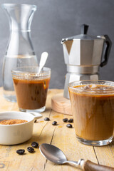 Close-up of two glasses with ice coffee, spoon, coffee beans, brown sugar with coffee pot and bottle of water, with selective focus, on wooden table, gray background, vertical