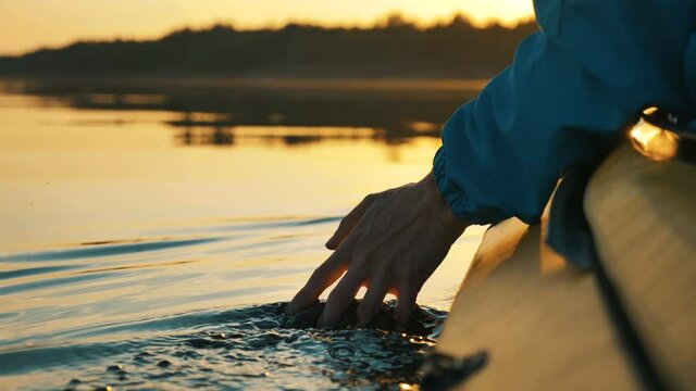 unity with nature, a man puts his hand into the water while sitting in a kayak against a golden sunset, camera slow motion close-up