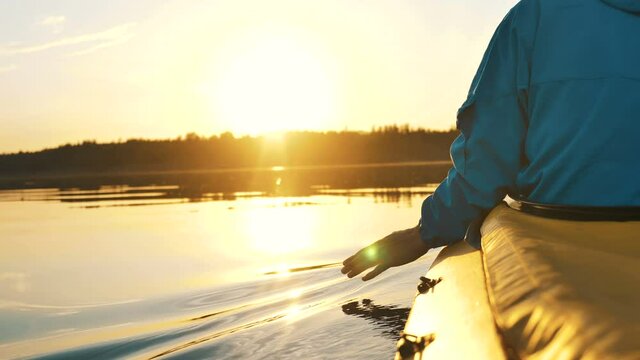 unity with nature, man runs hand through calm water while sitting in a kayak against a golden sunset, camera slow motion
