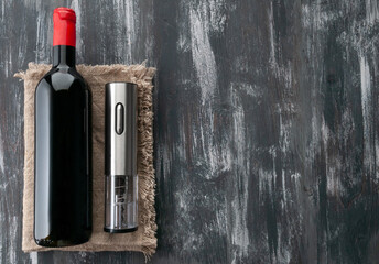 Gray electric corkscrew made of metal. Lies next to a bottle of wine on linen cloth. Dark wooden background. View from above.