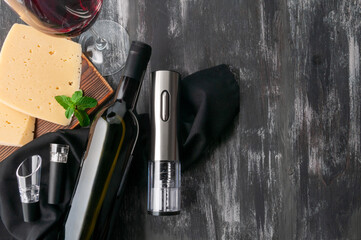 Gray electric corkscrew made of metal. Lies next to a bottle of wine, aerators and a vacuum stopper on black fabric. Next to a glass of wine and a piece of cheese. Dark wooden background. 