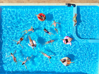 Aerial of friends having party in swimming pool with inflatable flamingo, swan, mattress. Happy...
