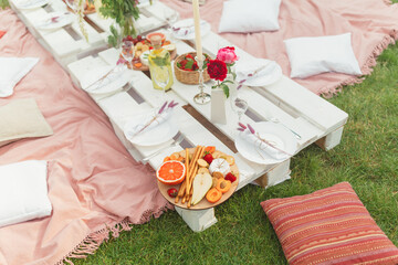 Festive summer picnic with delicious snacks, fruits and wine, decorated with beautiful flowers and candles in pink colors for hen party. Good food serving on white pallet table. Selective focus