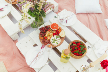 Delicious snacks, fruits, lemonade and wine on the white pallet table. Summer festive picnic decorated with beautiful flowers  for hen party. Good food serving. Seating on the wrap and pillows