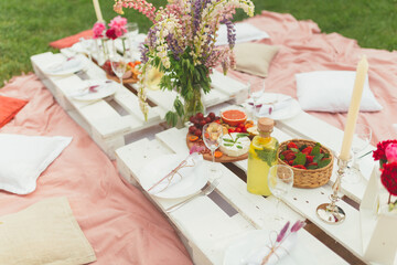 Summer picnic in garden with delicious snacks, fruits, lemonade and wine on white pallet tables. Decorated with beautiful flowers in pink colors for hen party. Seating on the wrap and pillows