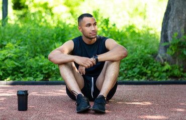 Handsome African American sportsman sitting on road, taking break after his training at park