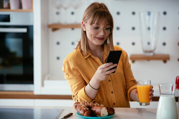 Beautiful woman drinking orange juice in the kitchen. Young woman reading the news online.	