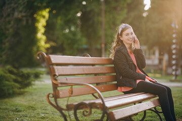 Young woman dressed in stylish wear having phone conversation with boyfriend. Discussing ideas for weekends during work break sitting outdoors on city bench.