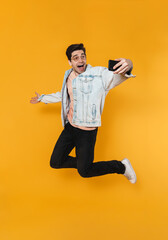 Photo of delighted caucasian man jumping while taking selfie on cellphone