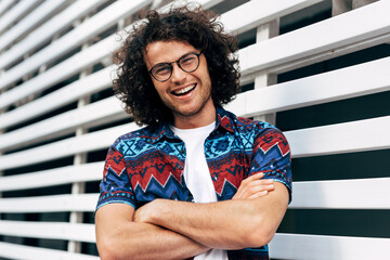 Portrait of stylish cheerful young man with curly hair smiling broadly with crossed arms in colroful shirt and eyewear. Happy male wearing casual trendy outfit enjoying the sunny summer day in town.