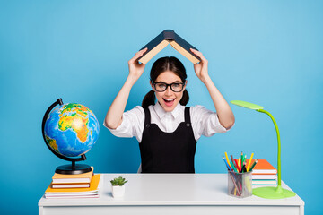 Photo of excited positive high school girl sit table put book above head have lesson break pause rest relax wear white blouse black overall uniform isolated blue color background