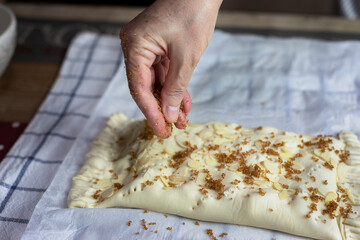 Female hand sprinkles brown sugar on a raw roll of puff pastry decorated with almond flakes.