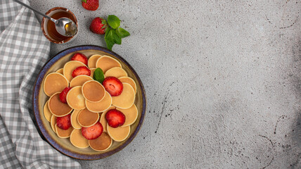 Tiny pancakes for breakfast. Cereal pancakes with strawberries and honey in plate on grey background. Trendy food.