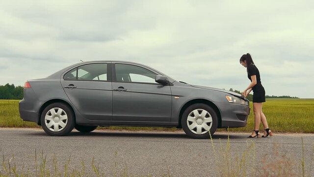 A young girl in a short dress approaches a broken car and opens the hood.The car suddenly broke down on a country road down while driving. Slow motion.