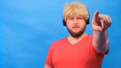 Naklejka premium freaky fat man in a wig and a pink T-shirt in headphones shows a finger on the camera at you against a blue background.