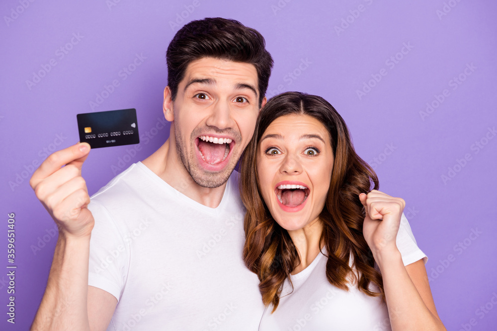 Poster Portrait of ecstatic two married people hold credit card win bank cashback service help raise fists scream yes wear white t-shirt isolated over violet color background