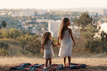Two little sisters in white clothes standing on a blanket watching the city while resting in nature