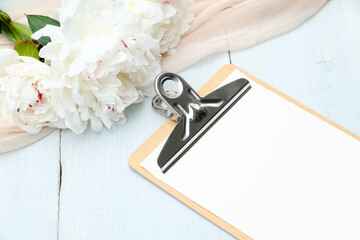 Paper clipboard mockup and peony flowers on blue wooden table. Flat lay, top view. Wedding planner, checklist, to do list concept.