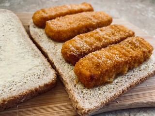 Closeup view of crispy fish fingers covered in salt and vinegar on buttered bread in the kitchen. Breaded fish finger sandwich preparation on wooden board.