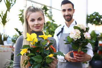 Charming young woman and bearded man in apron looking at camera and smiling while demonstrating flowers in plant store