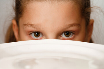Eyes of a girl peeking out from behind a white bowl of milk. Shallow depth of field. Focus  on the forehead and eyebrows