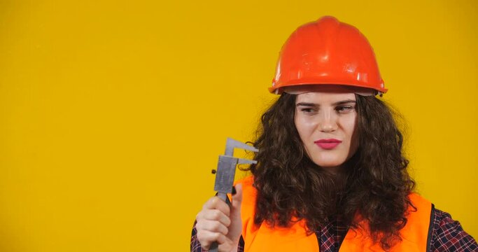 Attractive plus-size model posing. Curly-haired brunette on bright yellow studio background. Girl in red helmet and orange signal vest showing large size with caliper. Erotic Fantasy Concept