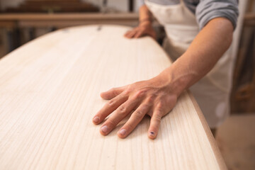 Caucasian male surfboard maker working in his studio, making a surfboard and inspecting it