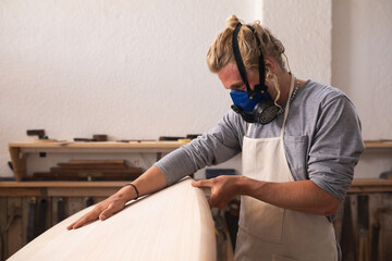 Caucasian male surfboard maker wearing a face mask working in his studio making a surfboard
