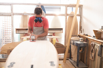 Caucasian male surfboard maker working in his studio with a protective apron and a breathing mask