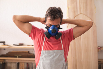 Caucasian male surfboard maker wearing a protective apron, putting on a breathing mask