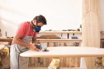 Caucasian male surfboard maker working in his studio, with a protective apron and a breathing mask