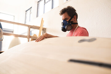 Caucasian male surfboard maker wearing a protective apron, inspecting a wooden surfboard