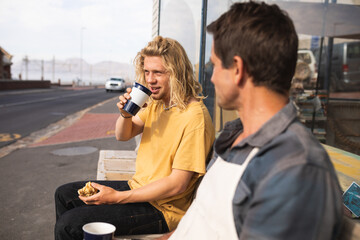 Two Caucasian male surfboard makers sitting on a bench and drinking coffee and having a snack