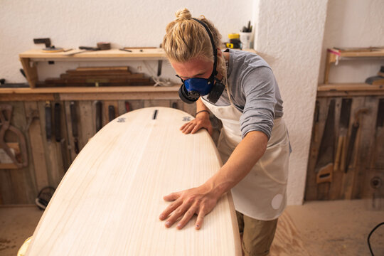 Caucasian male surfboard maker wearing a face mask, working in his studio making a surfboard