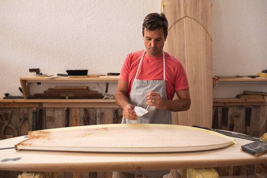 Caucasian male surfboard maker working in his studio, wearing a protective apron, putting on a mask