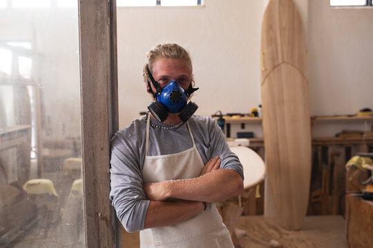 Caucasian male surfboard maker wearing a face mask, standing in his studio and looking at camera
