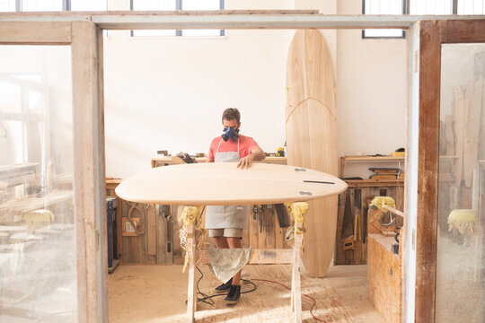 Caucasian male surfboard maker wearing a protective apron, inspecting a wooden surfboard