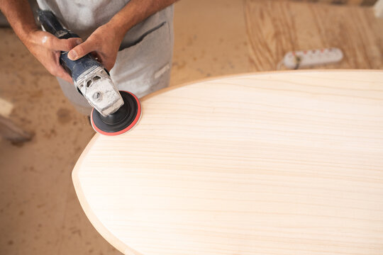 Caucasian male surfboard maker wearing a protective apron, shaping a wooden surfboard with a sander