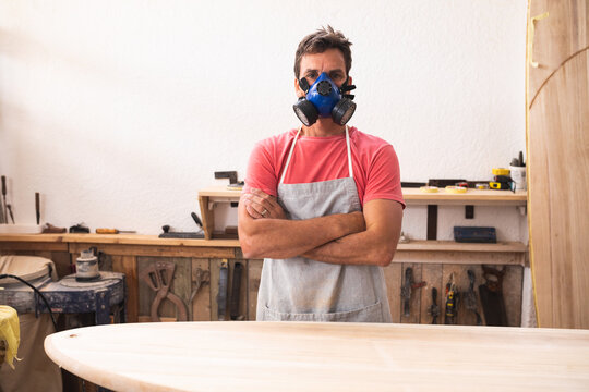 Caucasian male surfboard maker wearing a protective apron and a mask, looking at camera