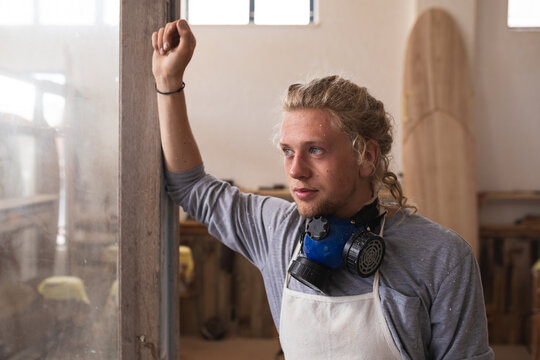 Caucasian male surfboard maker wearing a face mask, standing in his studio leaning on a door frame 