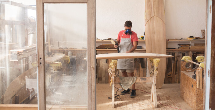 Caucasian male surfboard maker wearing a protective apron, inspecting a wooden surfboard - Powered by Adobe