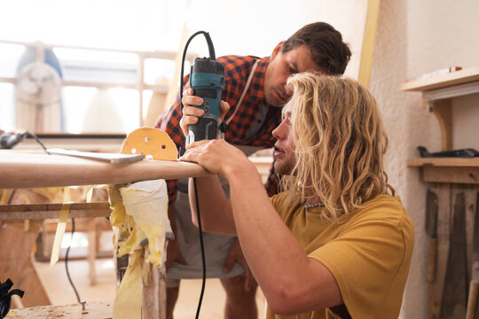 Two Caucasian male surfboard makers making a wooden surfboard together with a sander - Powered by Adobe