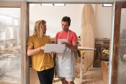 Two Caucasian male surfboard makers standing by the door frame and working on projects
