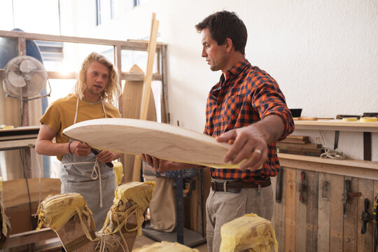 Two Caucasian male surfboard makers making a wooden surfboard together and inspecting it